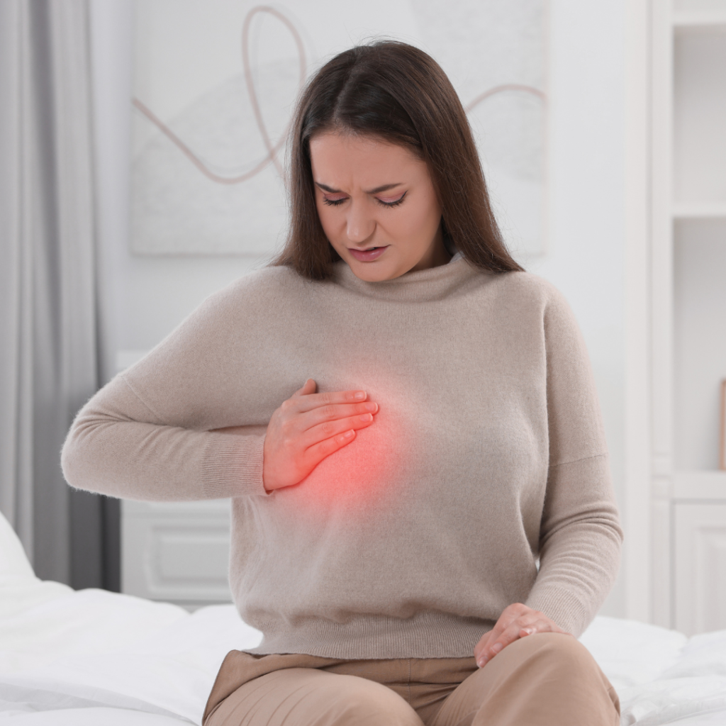A woman sitting on a bed holds her chest with a highlighted red area, symbolizing breast pain or discomfort.